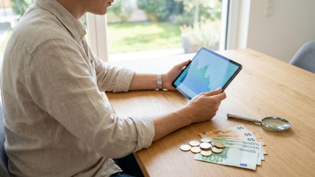 Un homme examine un graphique financier sur une tablette près de billets d'euros et d'une loupe sur une table en bois.